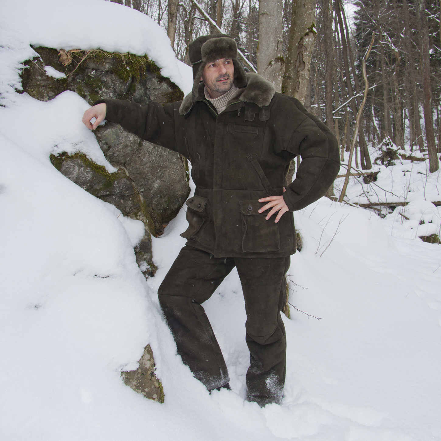 Model trägt Ansitzkleidung aus Lammfell. Jacke, Hose und Mütze. Stimmungsbild im verschneiten Wald.